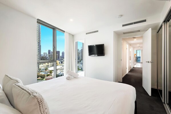 A hotel room with a large bed, a TV mounted on the wall, and a view of the cityscape through large windows.