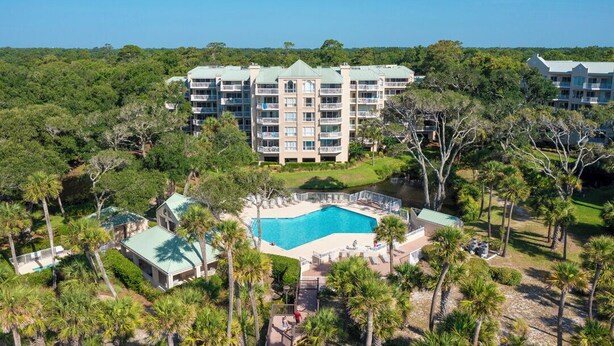 Um resort com piscina central, rodeado por palmeiras e um edifício grande com várias varandas.