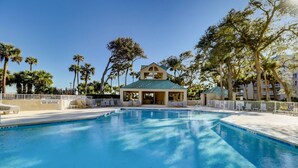 Uma piscina com espreguiçadeiras, um pequeno edifício com telhado verde e palmeiras.