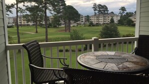 Un balcon agrémenté de fauteuils en osier et d’une table en pierre, offrant une vue sur un terrain de golf et des immeubles résidentiels.