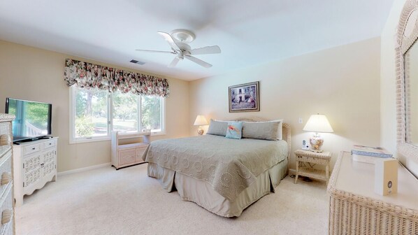 A bedroom with a bed, two bedside tables, a television, a window with curtains, a ceiling fan, and a framed picture on the wall.