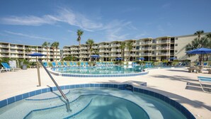 Una piscina con azulejos azules, rodeada de sillas de descanso y sombrillas, con un edificio de varios pisos al fondo.