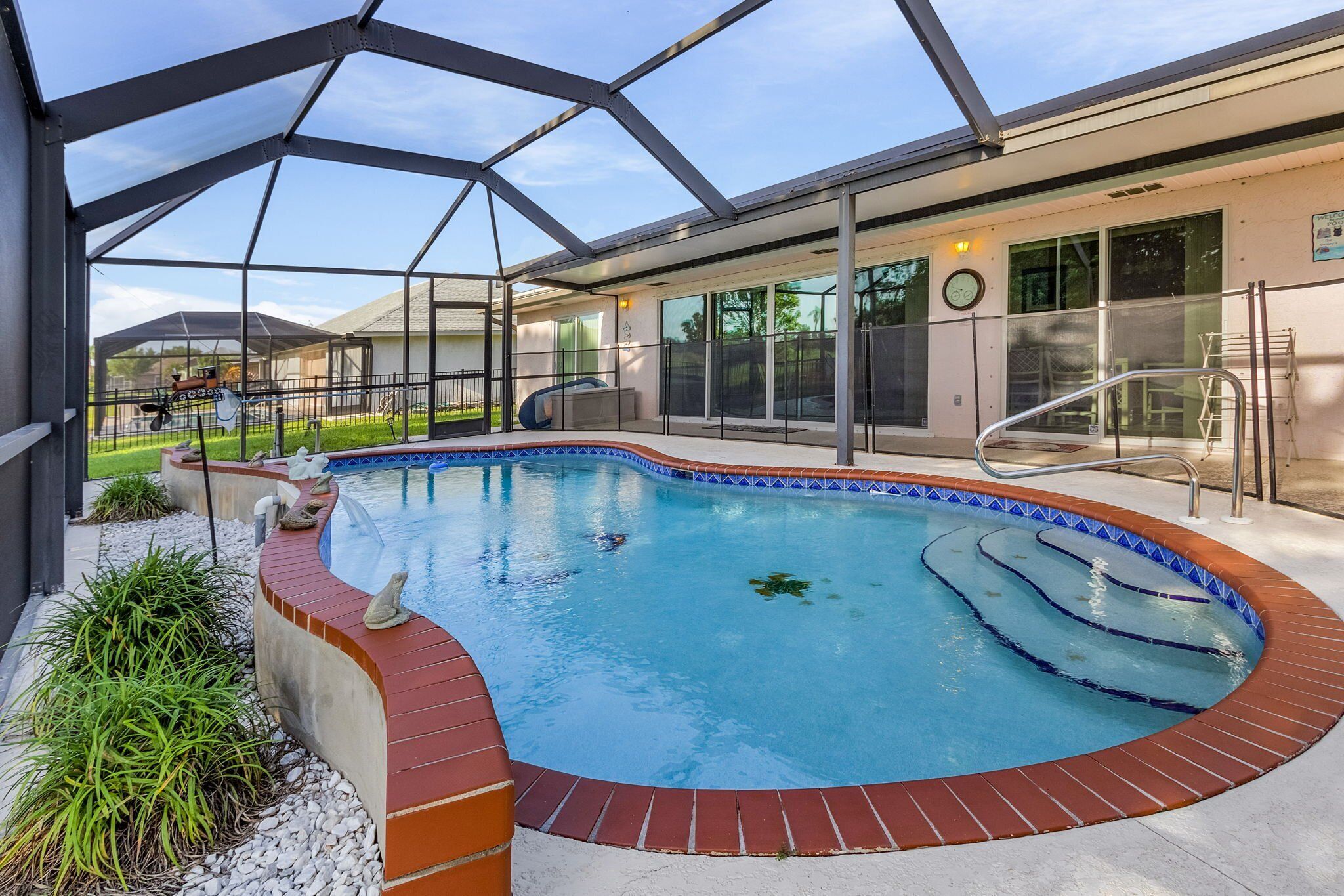 A swimming pool with a glass enclosure, surrounded by a brick border, and a covered patio area with a metal railing.