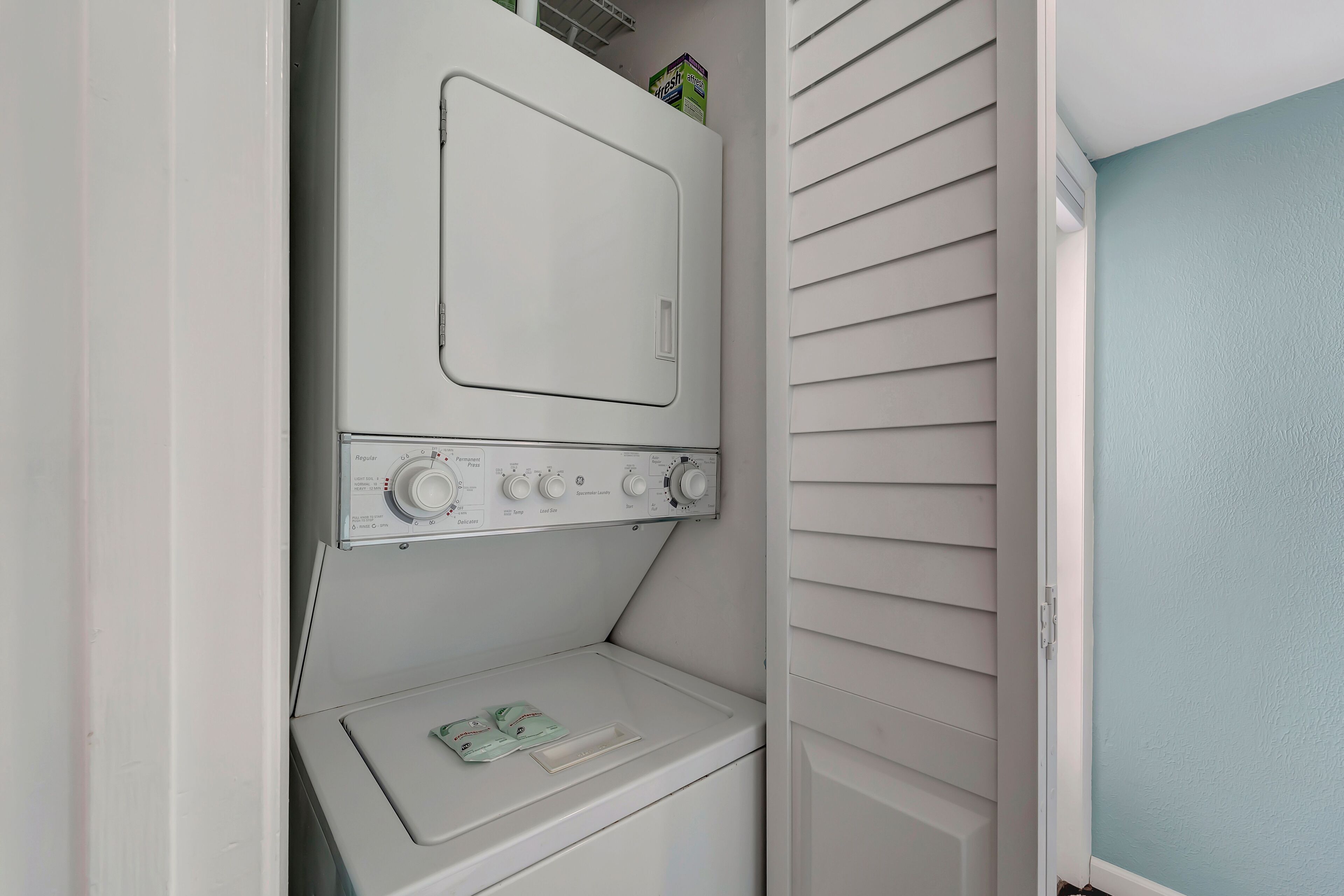 Een wasserette met een wasmachine en droger boven elkaar, een klein plankje met waszakjes en een deur naar een kamer met lichtblauwe muren.