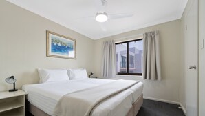 A hotel room with a bed, a bedside table, a ceiling fan, a window with curtains, and a painting on the wall.
