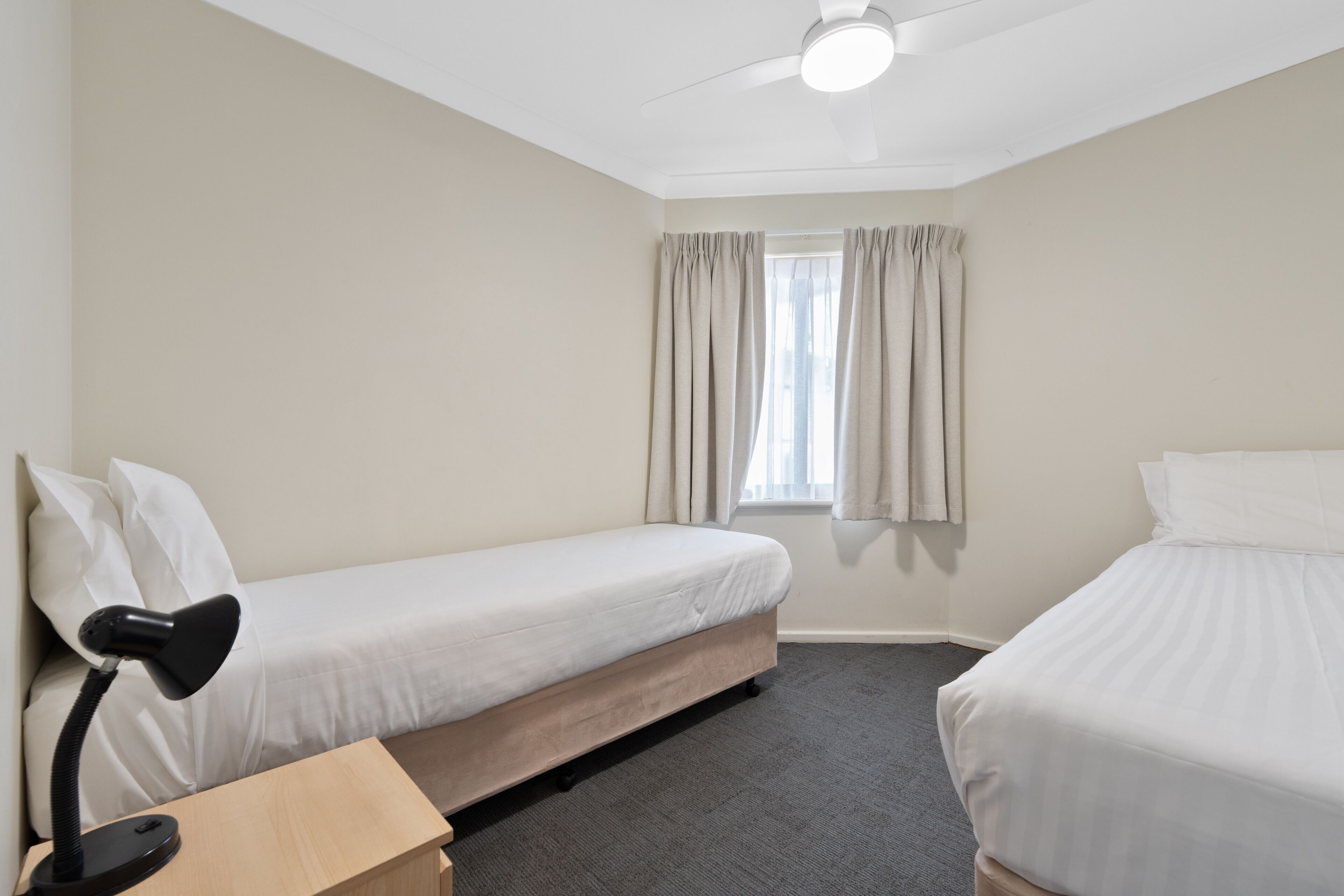 Une chambre d’hôtel avec deux lits simples, un bureau avec une lampe, un ventilateur de plafond et une fenêtre avec des rideaux.