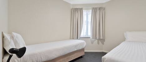 Une chambre d’hôtel avec deux lits simples, un bureau avec une lampe, un ventilateur de plafond et une fenêtre avec des rideaux.