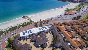 An aerial view of a coastal area with a large building featuring solar panels, surrounded by residential houses with red-tiled roofs, a beach, and a road with parked cars.