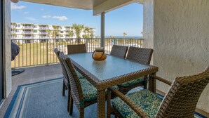A balcony with wicker furniture, a glass table, and a view of a building and palm trees. - CRC 1206 - Newly Updated Condo with Ocean and Pool View (St Augustine)