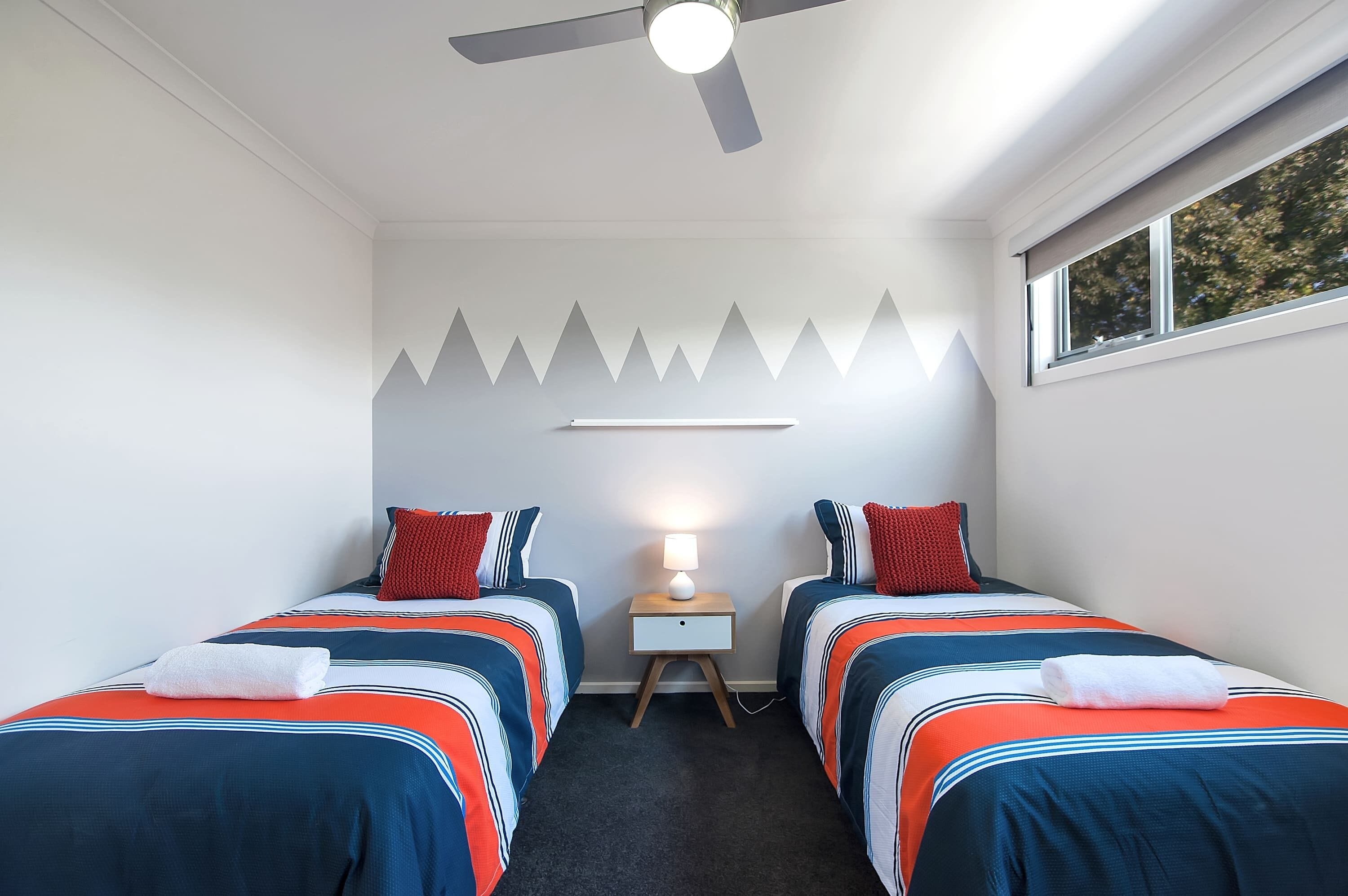 A room with two beds, a ceiling fan, a nightstand with a lamp, and a window with a view of trees.