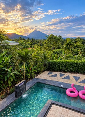 A swimming pool with a waterfall feature, surrounded by lush greenery and a view of a mountain. - Glass House Arenal Private Pool + Volcano Views (La Fortuna)