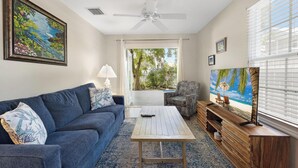 Un salon avec un canapé bleu, une table basse en bois, un téléviseur posé sur un meuble en bois et une grande fenêtre avec des stores.