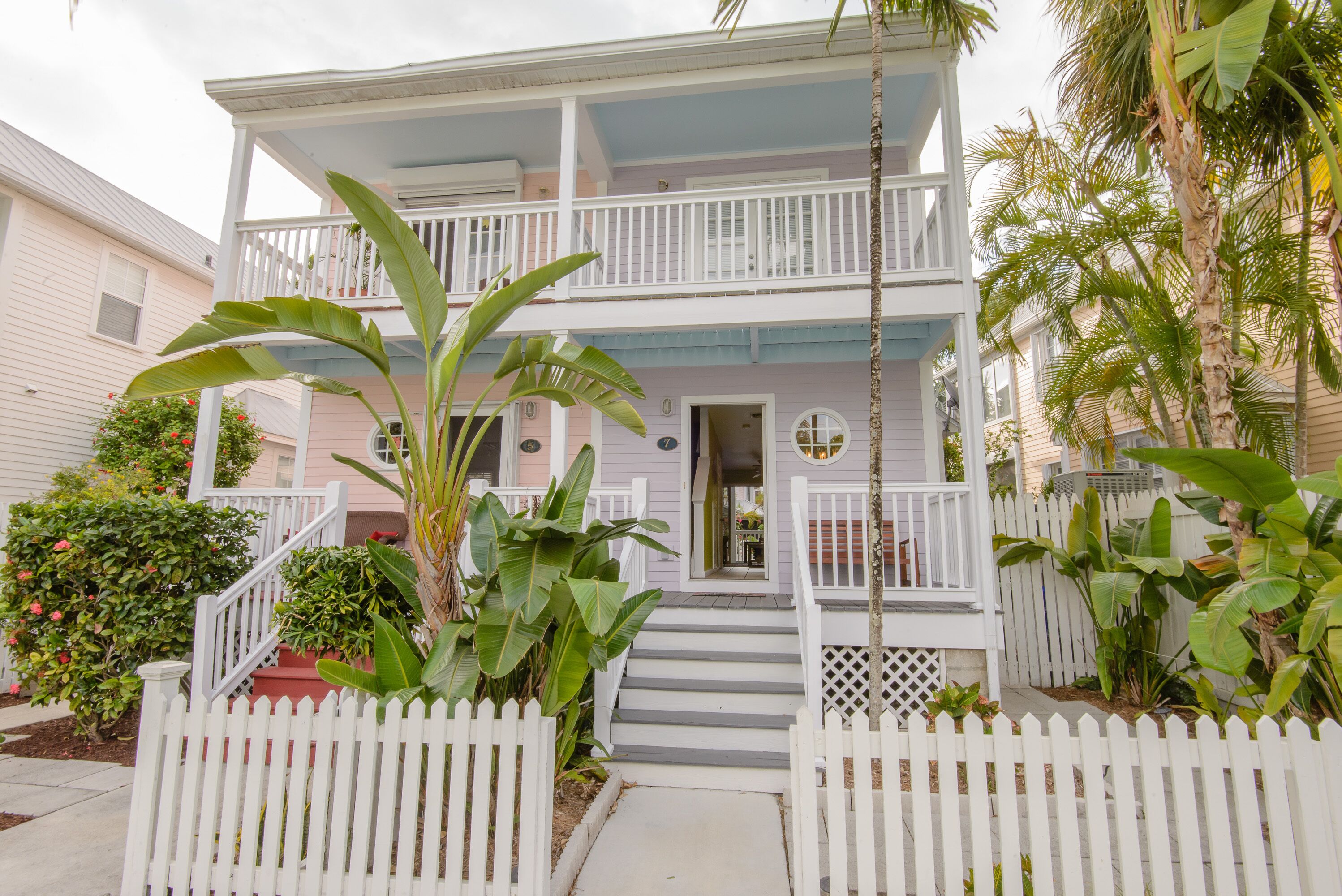 Une maison à deux étages avec un porche à l’avant, une clôture en bois peinte en blanc et des plantes tropicales.