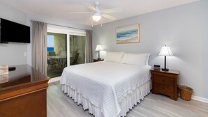 A bedroom with a bed, a desk, a television, a ceiling fan, and a view of the outdoors.
