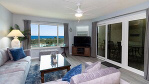 A living room with a large window, a ceiling fan, a flat-screen TV, and a sliding glass door leading to an outdoor area.