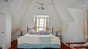 A bedroom with a sloped ceiling, a bed with a white and blue bedspread, two bedside tables with lamps, a window with curtains, and a ceiling fan.