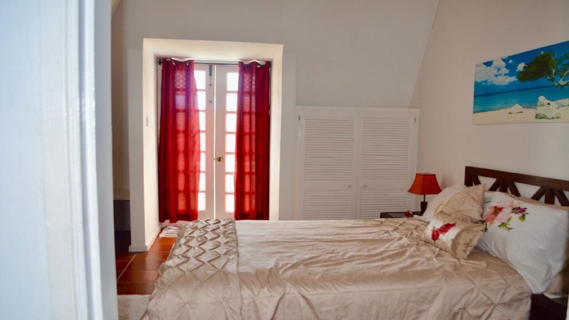 A bedroom with a bed, a nightstand, a lamp, a door with red curtains, and a painting on the wall.