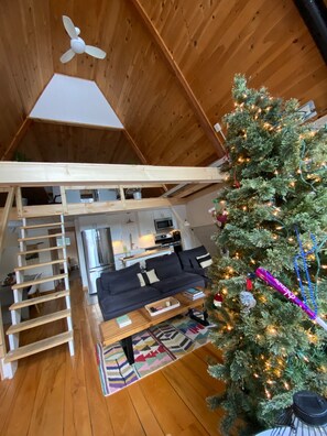 A cozy loft-style room with a wooden ceiling, a staircase, a kitchen area, and a Christmas tree. - Mod A Frame Cabin in the Woods with Hot Tub -close to Mt. Snow & Stratton! (Wardsboro)
