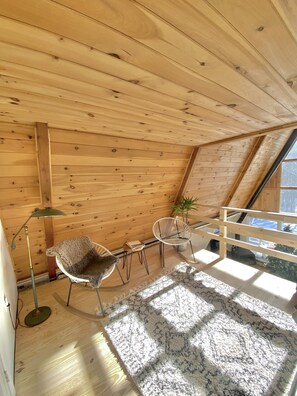 A cozy attic room with wooden walls, a sloped ceiling, a small table with chairs, a floor lamp, and a view through a window. - Mod A Frame Cabin in the Woods with Hot Tub -close to Mt. Snow & Stratton! (Wardsboro)