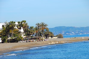 A beach with palm trees, a café, and a view of the sea and distant mountains.
