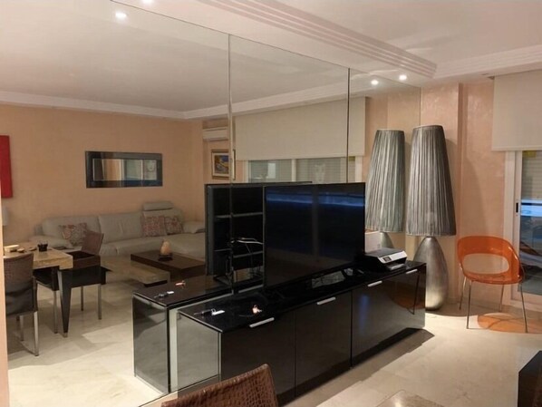 A modern living room with a large flat-screen TV, a sofa, a coffee table, and a dining area with a chair and table.