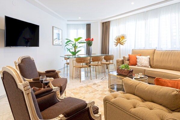 A modern living room with a sofa, armchairs, a glass dining table, and a flat-screen TV mounted on the wall.