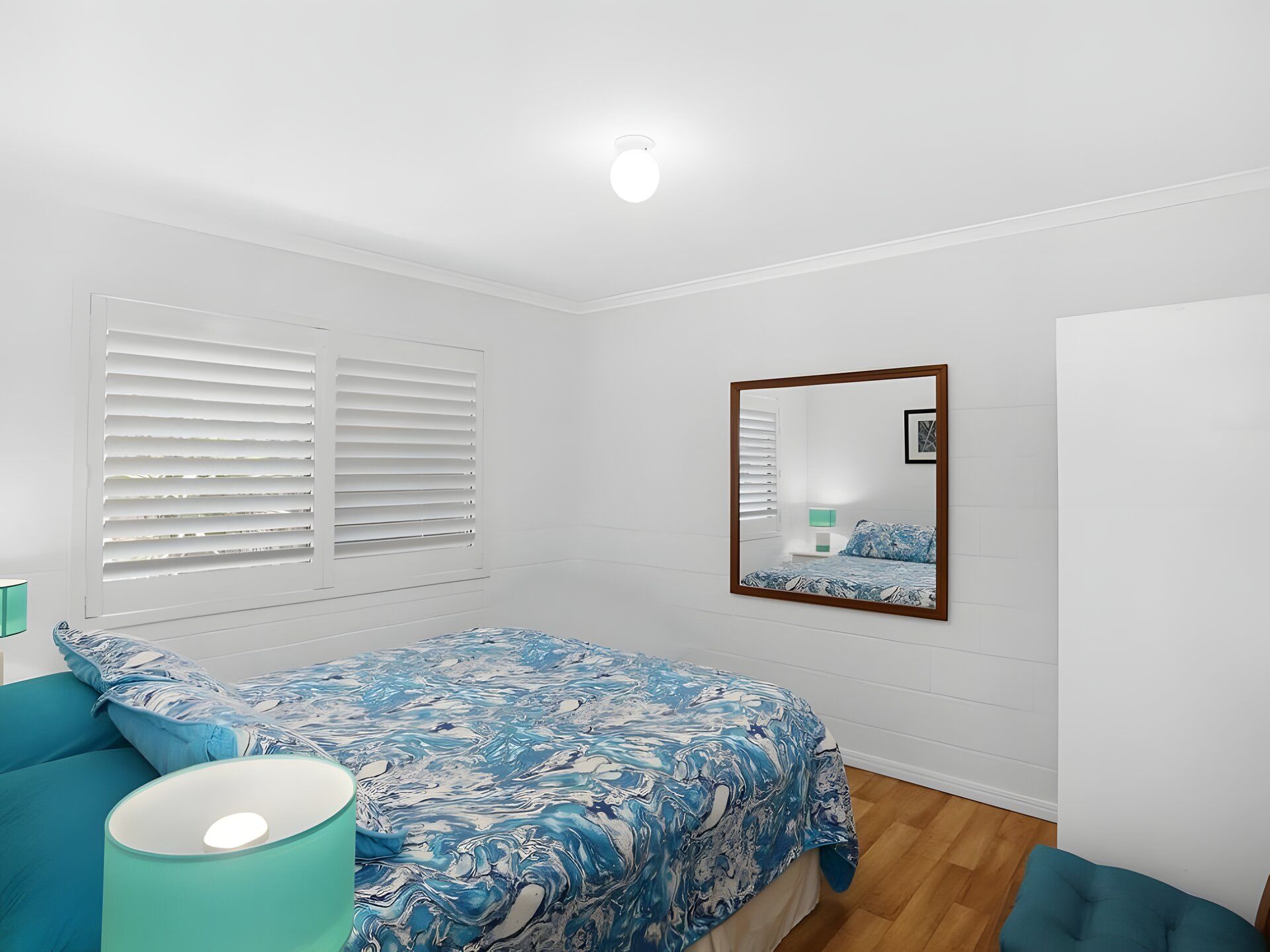 A bedroom with a bed, a bedside lamp, a mirror, and a window with blinds.