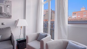 Une pièce moderne avec deux canapés, une table d’appoint en verre, une lampe et une vue sur les immeubles à travers de grandes fenêtres.