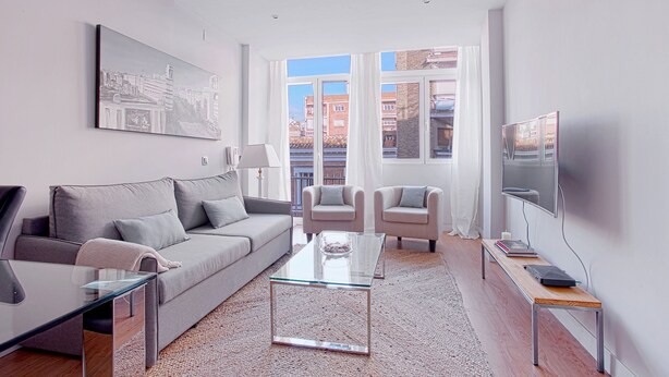 Un salon moderne avec un canapé gris, deux fauteuils, une table basse en verre, un téléviseur posé sur un meuble et une grande fenêtre donnant sur la ville.