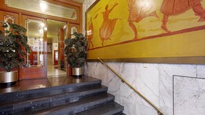 Un escalier en marbre avec une rampe dorée mène à un hall d’entrée agrémenté de boiseries et orné d’une grande fresque murale jaune.