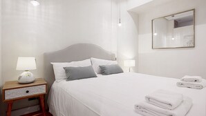 Un lit bien fait, avec du linge de lit blanc, deux lampes de chevet, une table de chevet en bois avec un tiroir et une serviette blanche pliée posée sur le lit.