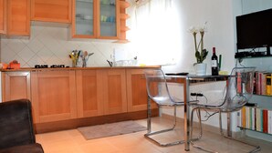 Ett kök med träskåp, en matplats med genomskinliga stolar, en tv och en bokhylla.