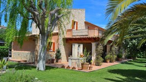 Un edificio de piedra con balcón, rodeado de un jardín bien cuidado con diversas plantas y una palmera.