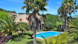 Una piscina rodeada de abundante vegetación y palmeras, con un edificio de piedra al fondo.