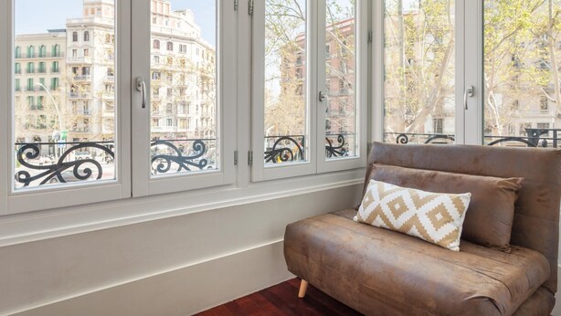 A room with a brown sofa and a patterned pillow, large windows with a view of buildings and trees. - ART HOUSE CITY CENTRE UNIVERSITAT (BARCELONA)