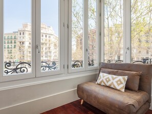 Una habitación con un sofá marrón y un cojín estampado, ventanas amplias con vistas a edificios y árboles.