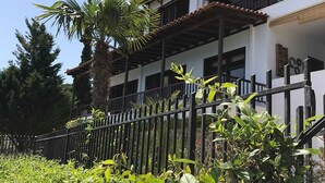 Um edifício de dois andares com fachada branca e telhado em telhas, cercado por uma cerca de metal preto e vegetação exuberante.