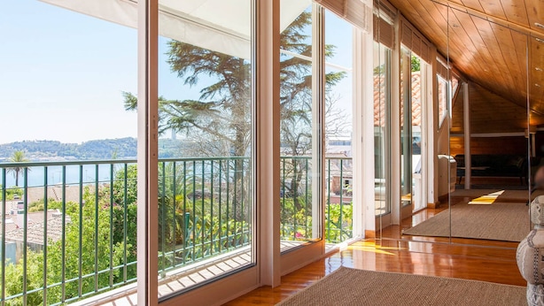 A spacious balcony with large glass doors, wooden flooring, and a view of greenery and distant hills. - Santos River View Terrace - Boutique Duplex (Lisboa)