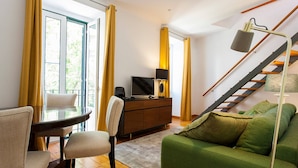 A modern living room with a green sofa, a glass table with chairs, a wooden cabinet with a TV, and a staircase with a lamp. - Santos River View Terrace - Boutique Duplex (Lisboa)