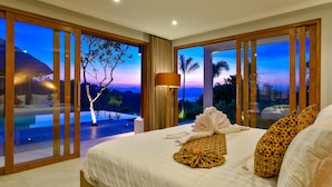 A spacious bedroom with a large bed, a view of the evening sky, and a balcony with a pool.