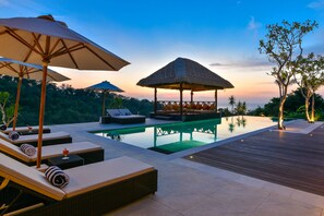 A serene poolside area with lounge chairs, umbrellas, and a thatched-roof structure.