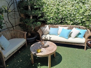 A wooden outdoor seating area with a round table and two wicker chairs, surrounded by greenery and a flowering bush.