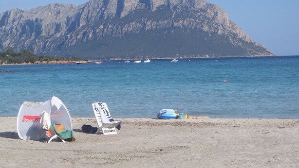 A beach with a large mountain in the background, a few boats on the water, and beach chairs and a tent on the sand.
