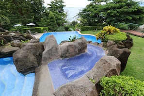 Une piscine agrémentée d’une cascade artificielle en roche, entourée d’une végétation luxuriante et offrant une vue sur la mer.