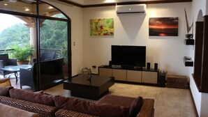 Ein Wohnzimmer mit einer Couch, einem Couchtisch, einem Fernseher und einem Balkon mit Stühlen und Pflanzen.