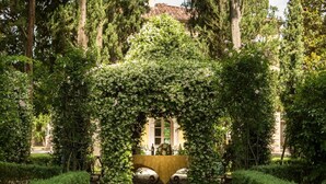 Un jardin avec une arche centrale recouverte de verdure, entouré d’arbres et avec un bâtiment en arrière-plan.