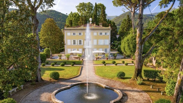 Un grand bâtiment avec une fontaine au centre, entouré d’un jardin bien entretenu, avec des arbustes taillés et un chemin de gravier menant au bâtiment.