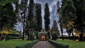 Un sentier de jardin mène à un bâtiment doté d’une arche centrale, entouré de grands arbres et de haies bien entretenues.