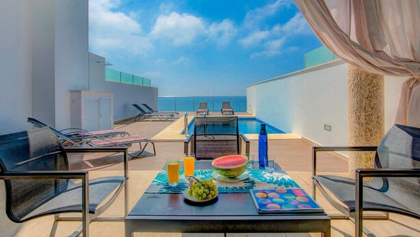 Une terrasse sur le toit avec une piscine, des chaises longues et une table où sont disposés une pastèque, des verres et un assortiment de fruits.