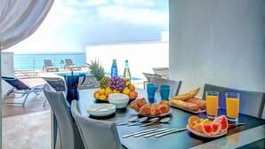 Une table de petit-déjeuner avec des fruits, des croissants et du jus d’orange, placée sur un balcon donnant sur l’océan.
